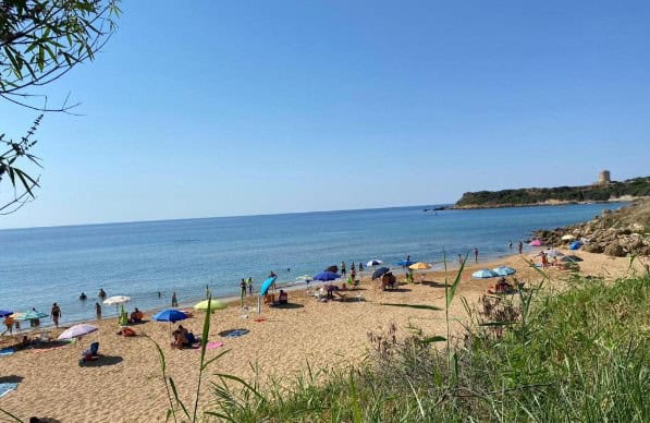 Peaceful sandy beach with people under beach umbrellas and the blue sea beyond with a cape in the distance to the right on a sunny day