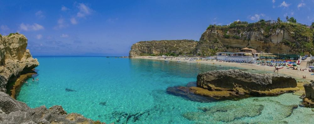 over water image of Baia di Riaci, with trransparent turquoise calm sea, white sandy beach with beachfront restauramt terrace and sheer cliffs behind. some people swimming and others on the beach under colourful umbrellas.