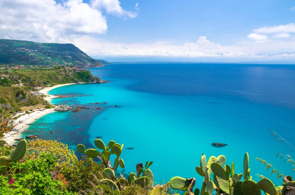 Capo Vaticano beaches aerial view from cliffs platform, sandy beach, green mountains, Sicilia island on horizon., blue sky white clouds background and cactus plants foreground