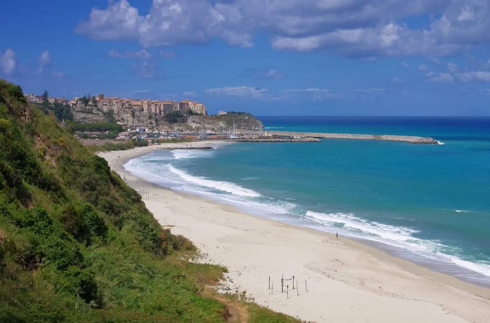 The long, wide white sand beach of Contura in Tropea, with small breaking waves on the shore and blue sea, on a sunny day with a few puffy clouds.On the edge of the beach is some green vegetation, and in the distance is the old town buildings of Tropea above the cliffs.