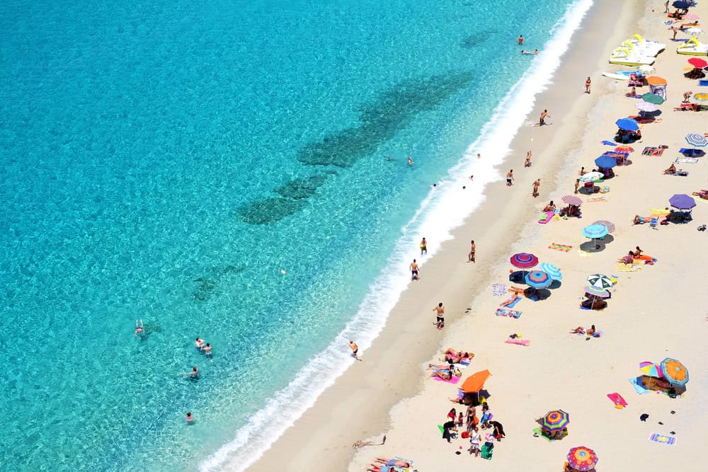 aerial view of Rotonda beach, one of the beaches in Tropea, with white sand beach and turquoise coloured sea with gentle waves lapping the shoreline. some people swimming and others under colourful umbrellas on the beach