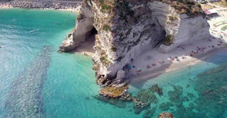 Aerial view of a large white rocky outrcop and sheer white cliffs with a triangular shaped cave like beach with white sand, surrounded by a clear blue sea, where the reefs below are visible. its a sunny day.