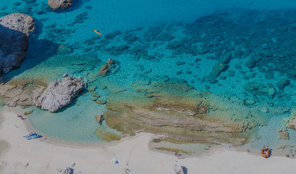 Aerial view of praia di fuoco beach, from the cliffs above. White sand beach with clear turquoise blue sea and reefs visible below. Two kayaks and one small inflatable boat on the shore with two people walking on the beach on a sunny day.