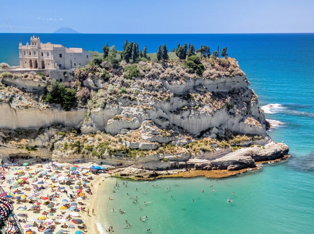 historic church of Santa maria della isola in Tropea on rocky outcrop with white sand beach and clear blue sea below on a sunny day. many colourful beach umbrellas and people swimming in the sea