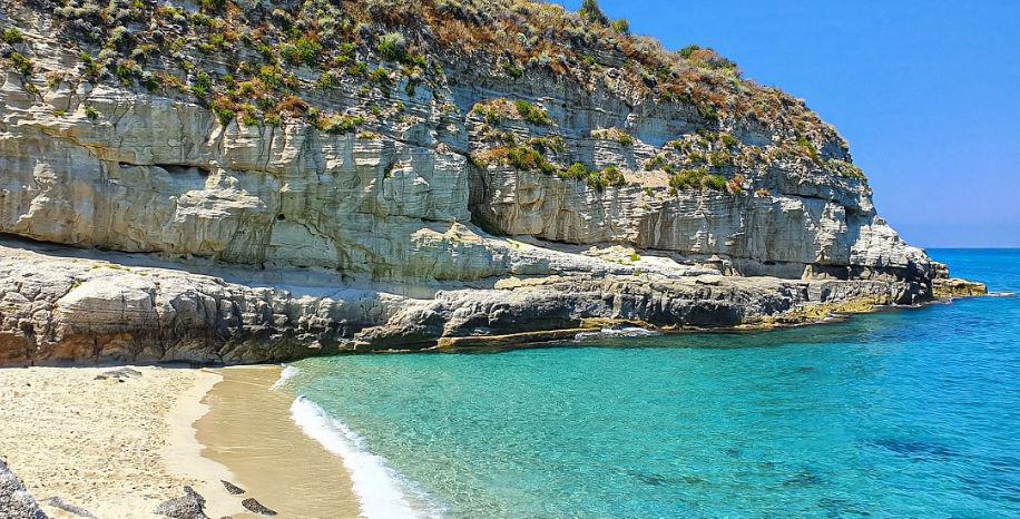 small white sand beach with small wavelets lapping the shore. A clear blue sea and white cliffs with green vegetation dispersed along the upper parts. the beach is deserted and it is a sunny day with clear blue skies.