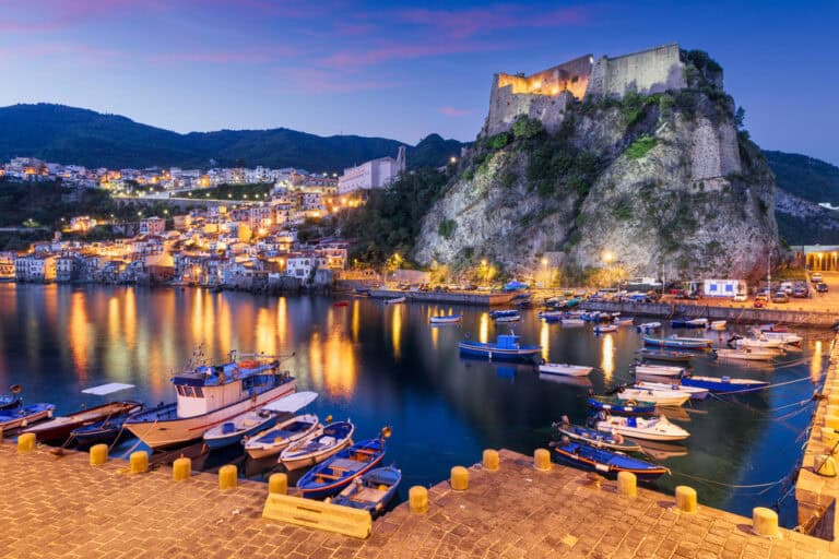 Scenic view of a harbor with boats and a historic castle at dusk in Southern Italy.