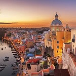 Sunset aerial view of procida harbour with pastel coloured houses and the harborfront promenade with moored boats.