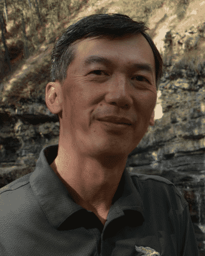 A smiling man outdoors with rocks and trees in the background.