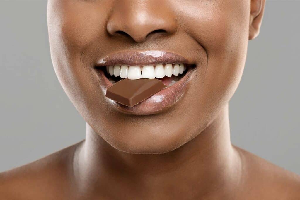 Smooth-skinned woman smiling with chocolate bar on lips, close-up portrait.