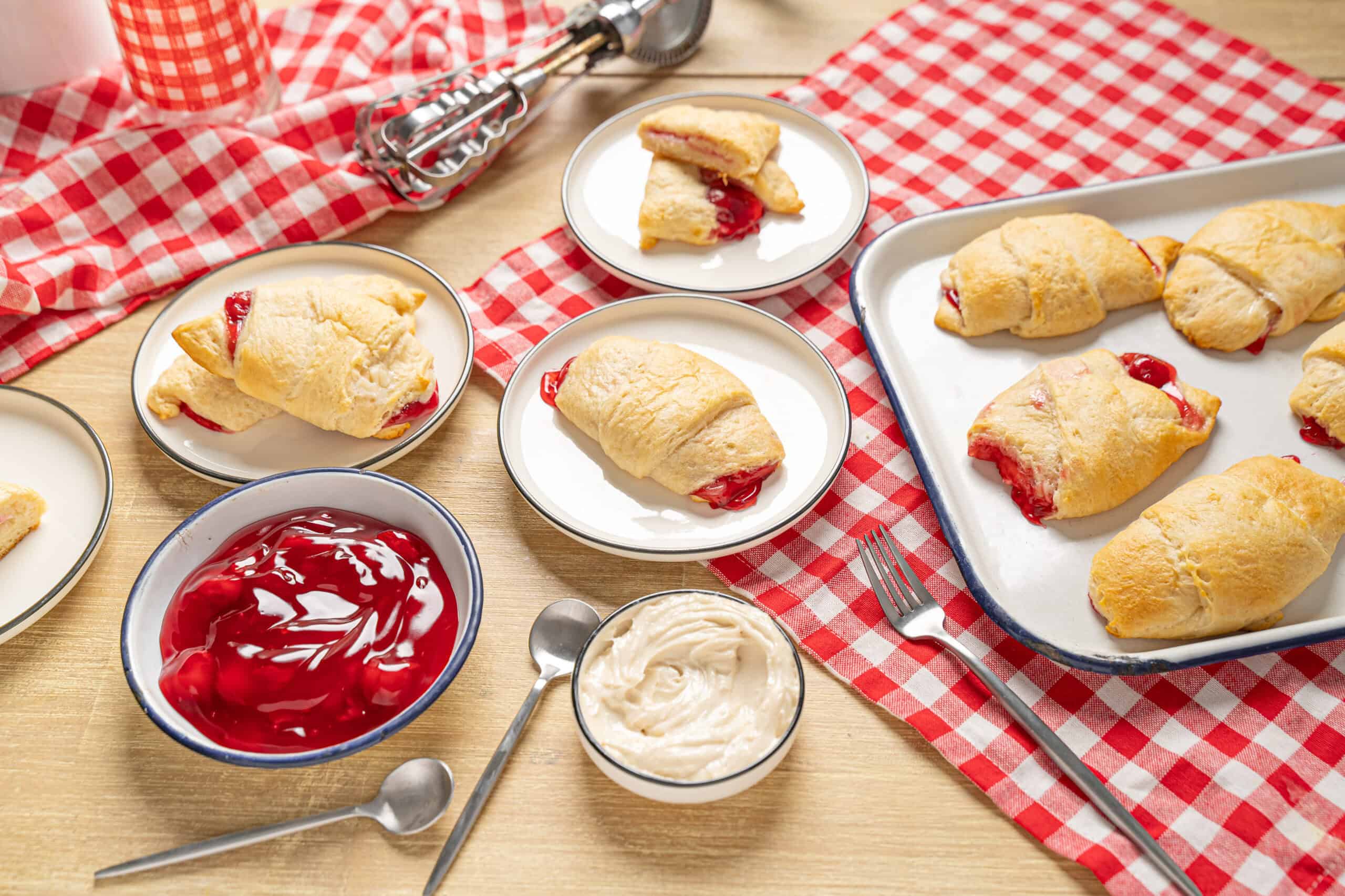 Soft homemade cherry turnovers on a baking sheet with bowls of cherry jam, whipped cream, and a fork, served on a checkered red and white tablecloth - delicious dessert recipe from Hey Recipe.