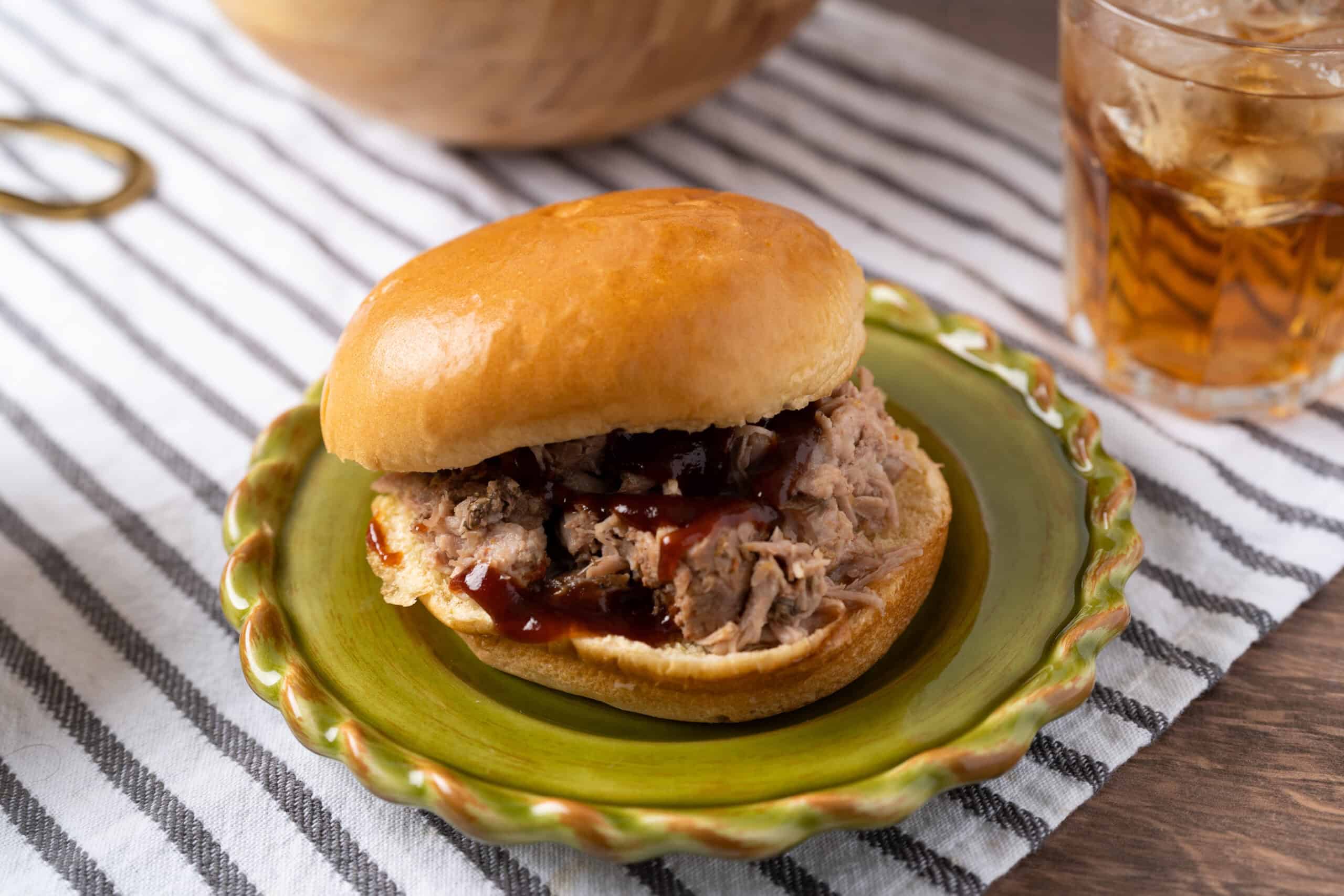 Savory pulled pork slider sandwich with barbecue sauce on a decorative green plate, served on a striped tablecloth with a glass of iced tea in the background.