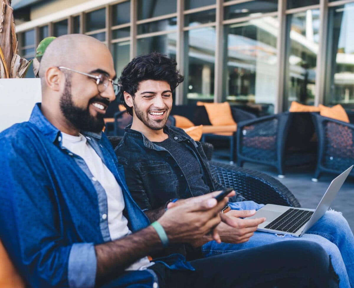 Modern happy ethnic men gathering on sofa in outside cafe and using mobile phone together while having discussing and laughing