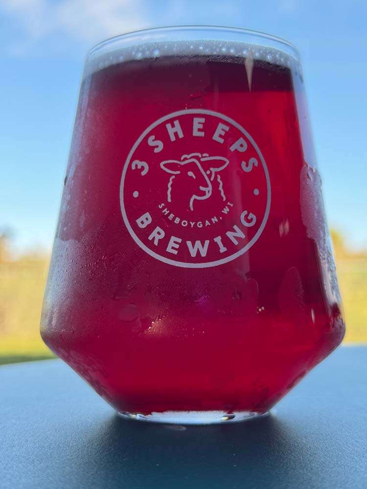 Rich red craft beer in a stemless glass with Sheep's Brewing logo, outdoors against a blue sky background.