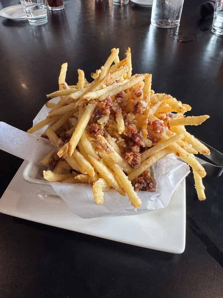 Crispy French fries topped with bacon bits on a white plate at a restaurant.