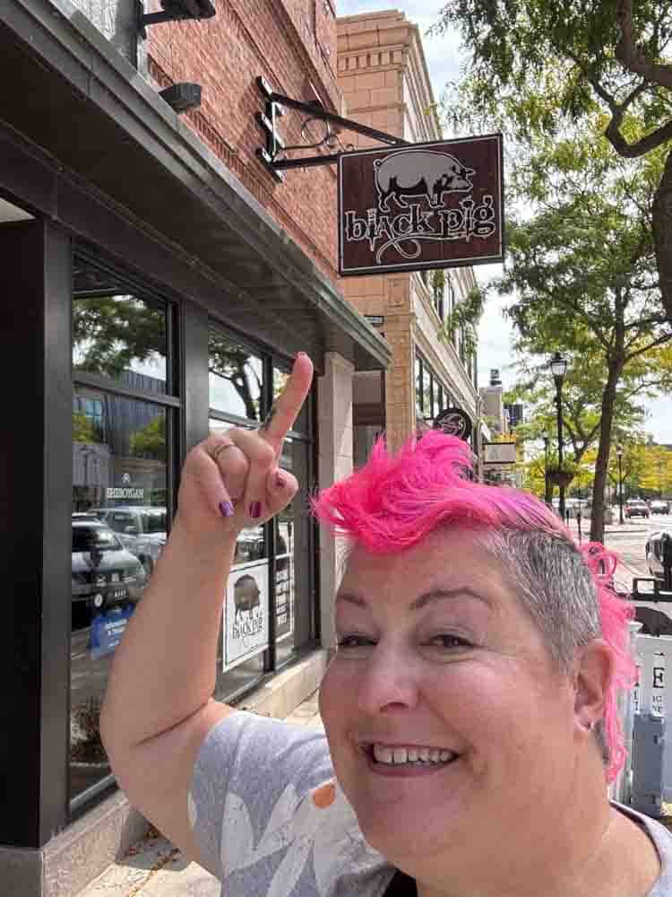 Black Pig restaurant sign outside storefront on city street, woman with vibrant pink hair pointing at sign, enjoying vibrant, lively atmosphere, trendy lifestyle, hip grandmother life, bold look.