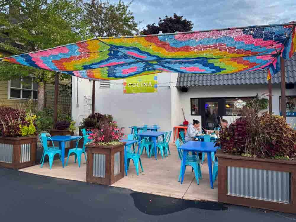 Colorful outdoor patio dining area at Hip Grandma Life restaurant with vibrant canopy, blue tables and chairs, and surrounding plants, enhancing the relaxed, welcoming atmosphere.