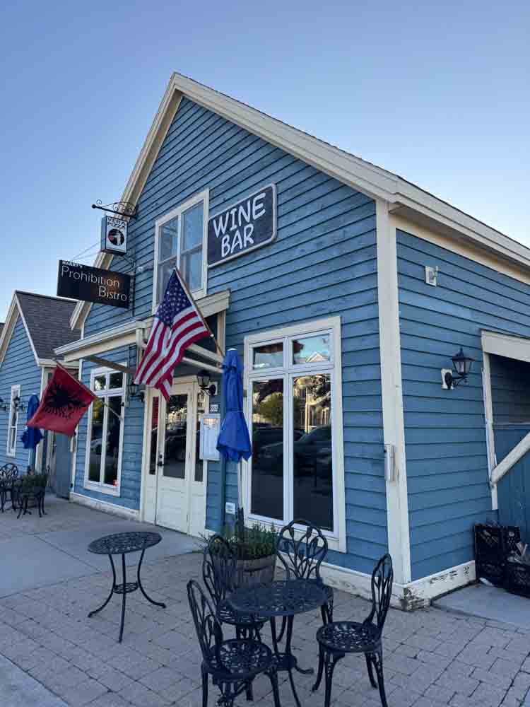 Cozy blue building with large windows, American flags, outdoor seating, and a "Wine Bar" sign, perfect for social gatherings or a nice evening out, as featured on Hip Grandma Life.