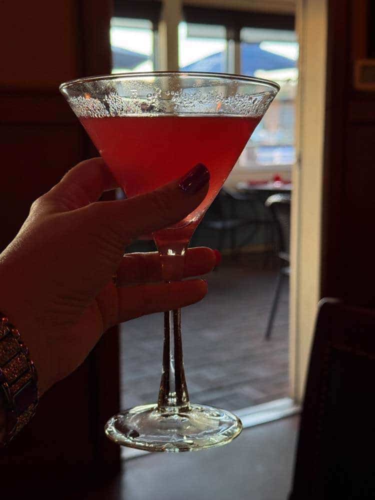 Red cocktail glass being held by a person's hand indoors, with a blurred background of a patio and windows, emphasizing a relaxing and stylish lifestyle for mature women.