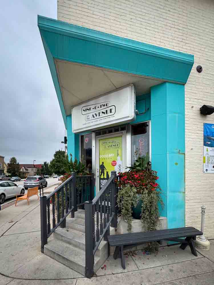 Bright turquoise storefront with stairs, plants, and a sign for 2nd Avenue; welcoming entrance to a podcast or radio station; vibrant decor and urban setting.