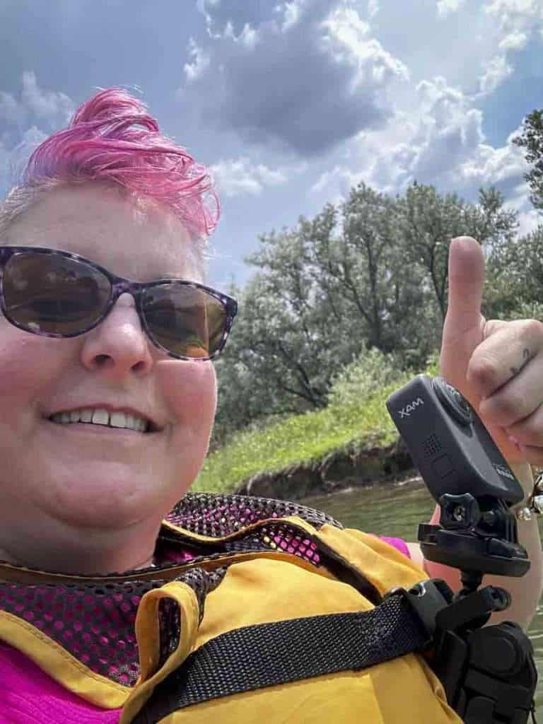 Bright pink-haired woman wearing sunglasses and life vest enjoying kayaking outdoors on a sunny day.