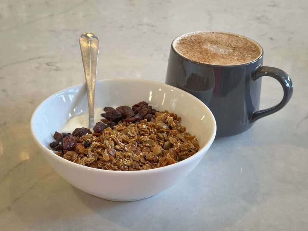 Nutritious breakfast bowl with granola, dried fruits, and dark chocolate accompanied by a hot cup of cocoa or coffee on a light-colored table.