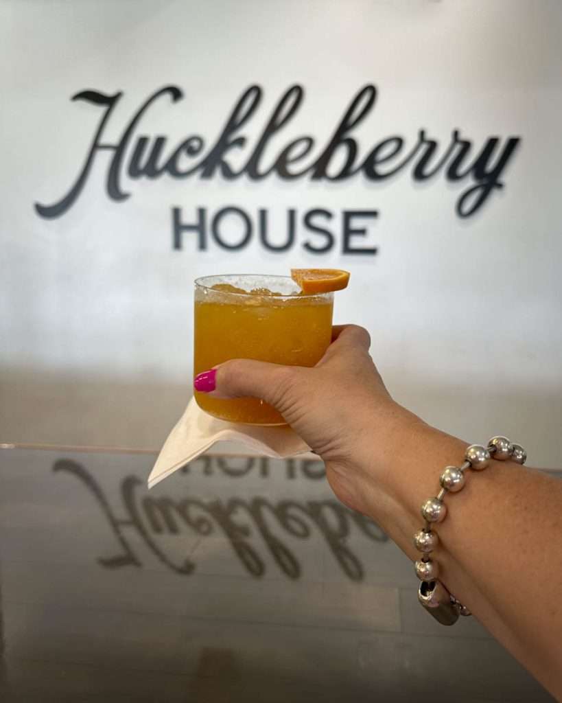 Refreshing orange cocktail with a garnish, held by a woman wearing a silver bead bracelet, in front of the Huckeberry House sign.