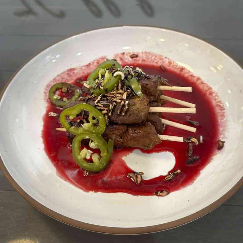 Juicy beef skewers topped with fresh jalapeños and seeds, served over a vibrant red sauce, garnished with sunflower seeds, perfect for flavorful, spicy outdoor dining.