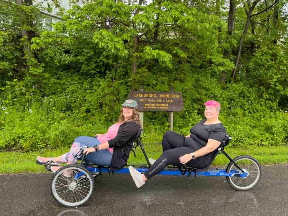 Women riding a tandem recumbent tricycle in front of a green wooded area in Minnesota, enjoying outdoor adventure and active lifestyles.