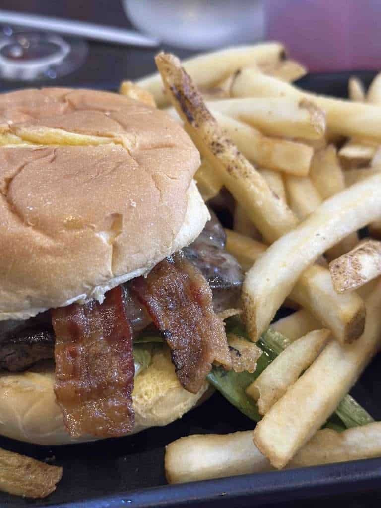 Bacon cheeseburger with lettuce and tomato served with crispy French fries on a black tray.