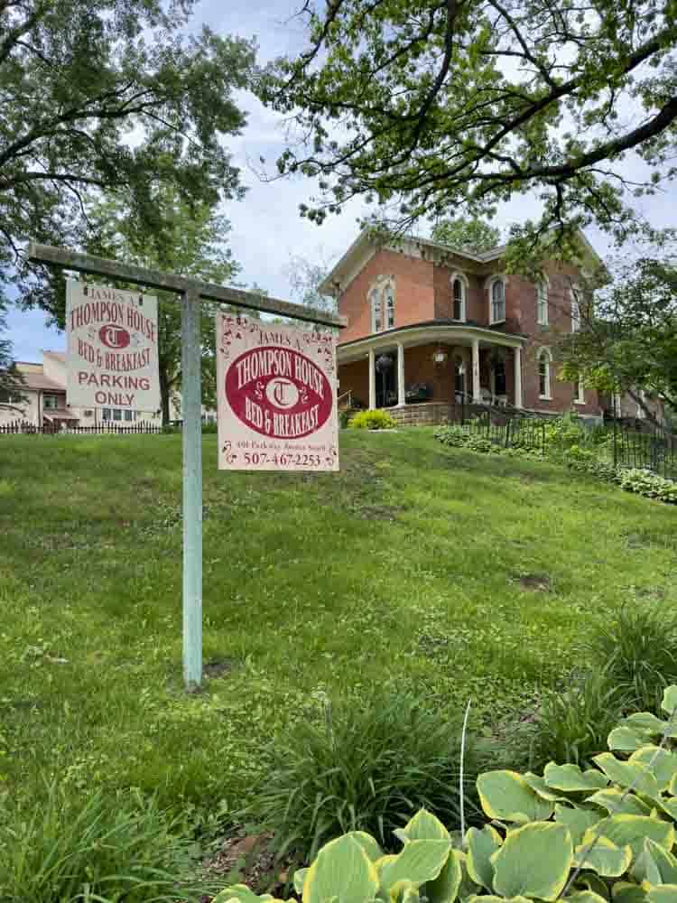 Thompson House Bed & Breakfast sign in front of a historic brick house surrounded by lush greenery and trees in a scenic neighborhood.