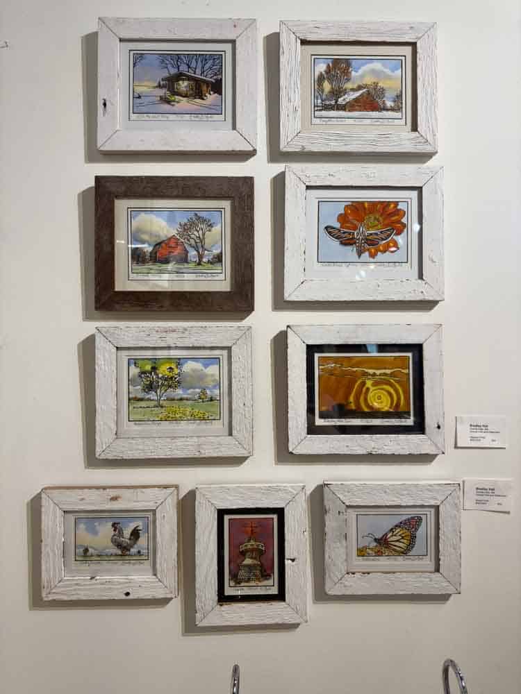 Colorful collection of framed artwork featuring rustic landscapes, butterflies, animals, and vibrant scenes, displayed on a white wall as part of an art exhibit celebrating nature and rural life.