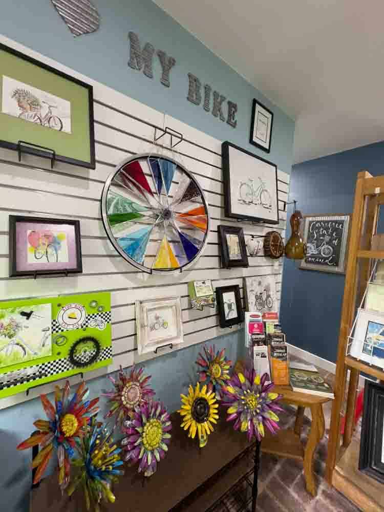 Colorful wall decor with framed artwork and decorative bicycle wheel, inspiring art pieces, and a display of vibrant sunflower and floral metal sculptures at Hip Grandma Life shop.