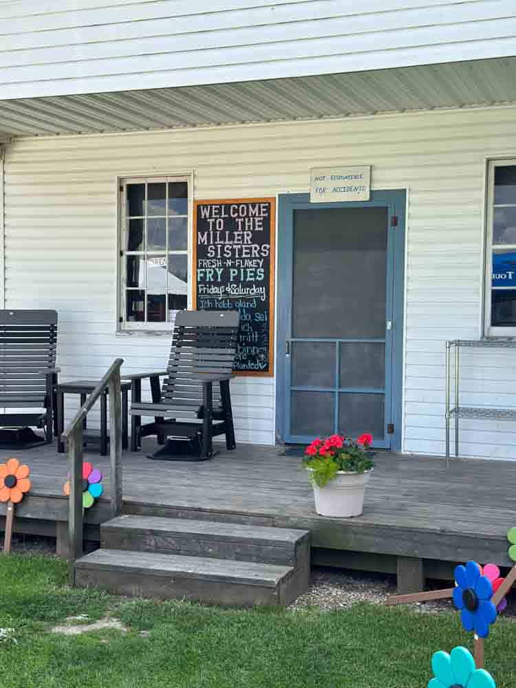 Charming porch with seating, decorative flowers, and a welcoming sign at the Miller Sisters farm stand, offering fresh fry pies and homemade treats, perfect for a cozy rural getaway.