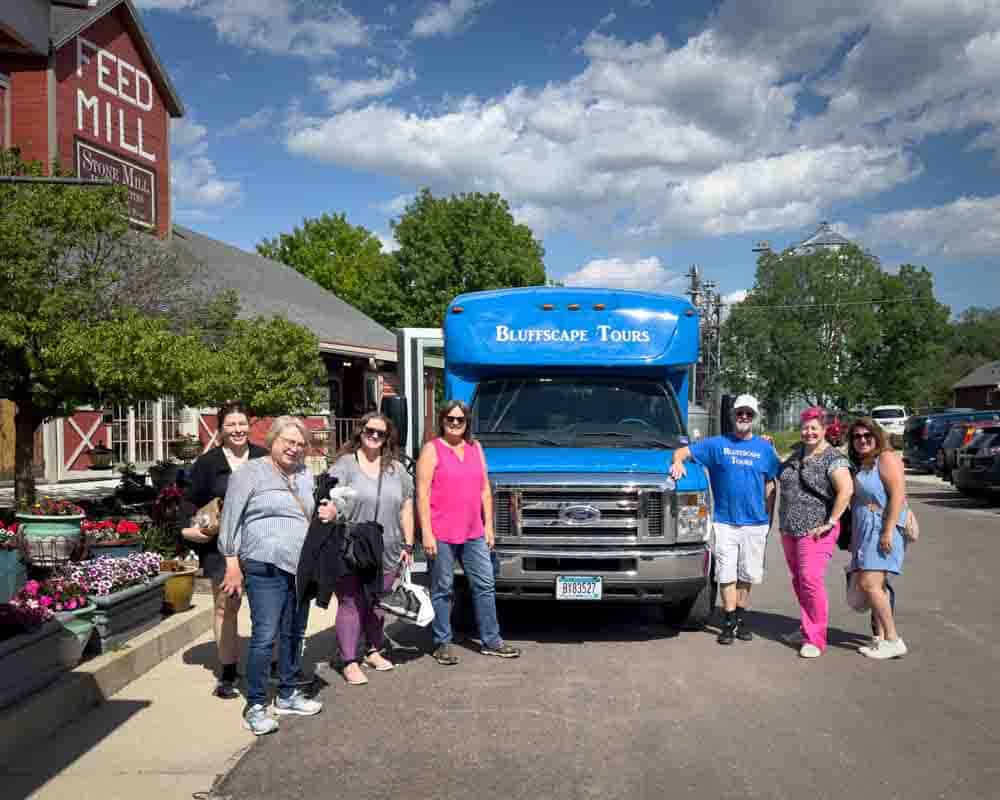 Colorful group of women and a man enjoying a scenic tour with Bluffscape Tours in front of a tour bus in a charming town setting.