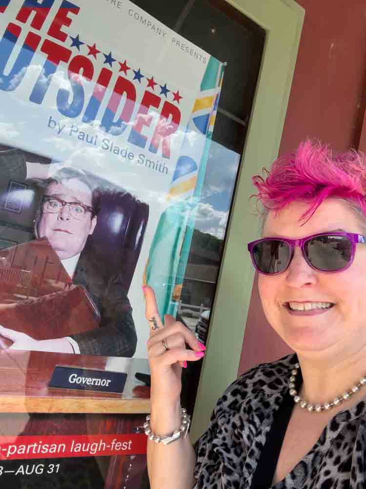 Bright pink-haired woman wearing sunglasses and jewelry pointing at a governor campaign poster outside, promoting humor and lively personality, perfect for lively political and social engagement.