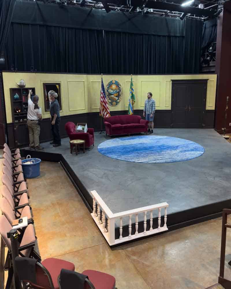 Flags and stage setup featuring American and state flags, a red velvet sofa, and a blue rug in a theater or community center for a political or public event.
