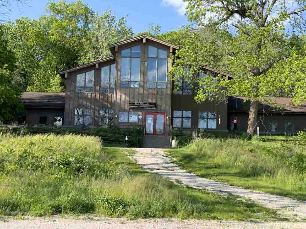 Rustic woodland-style building with large windows and a red door, surrounded by lush trees and greenery, ideal for outdoor activities and nature retreats.