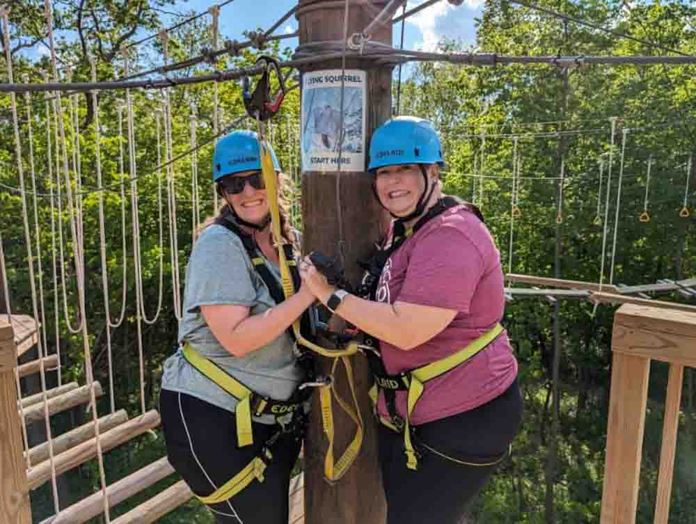 Happy women in safety harnesses and helmets on outdoor treetop adventure park, enjoying ziplining and nature activities.