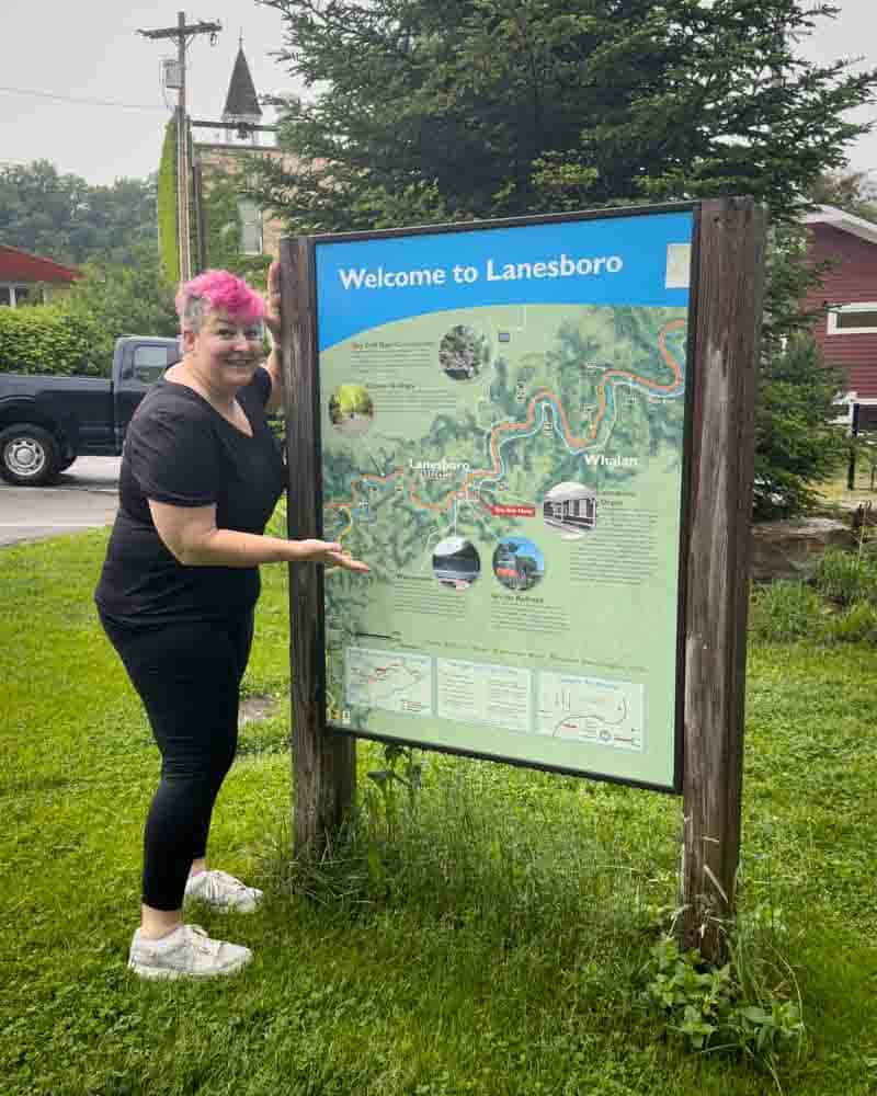 Vibrant woman with pink hair pointing at a welcome sign for Lanesboro, Minnesota, surrounded by lush greenery and trees.