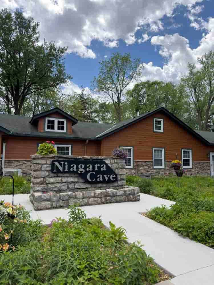 Charming rustic house with a stone and wood exterior, surrounded by lush greenery and flowering plants, under a partly cloudy sky, labeled Niagara Cave for a scenic outdoor experience.