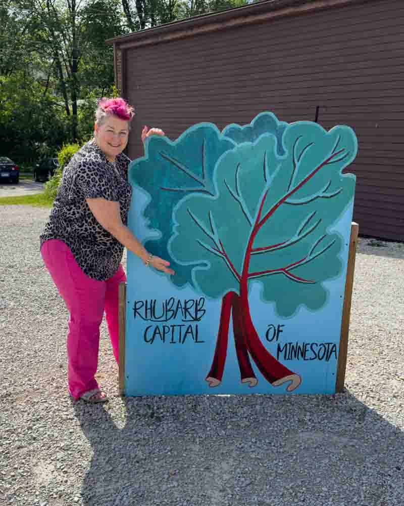 Rhubarb capital of Minnesota sign, vibrant woman outdoors holding rhubarb-themed artwork, celebrating local agriculture, rural life, and community in Minnesota.