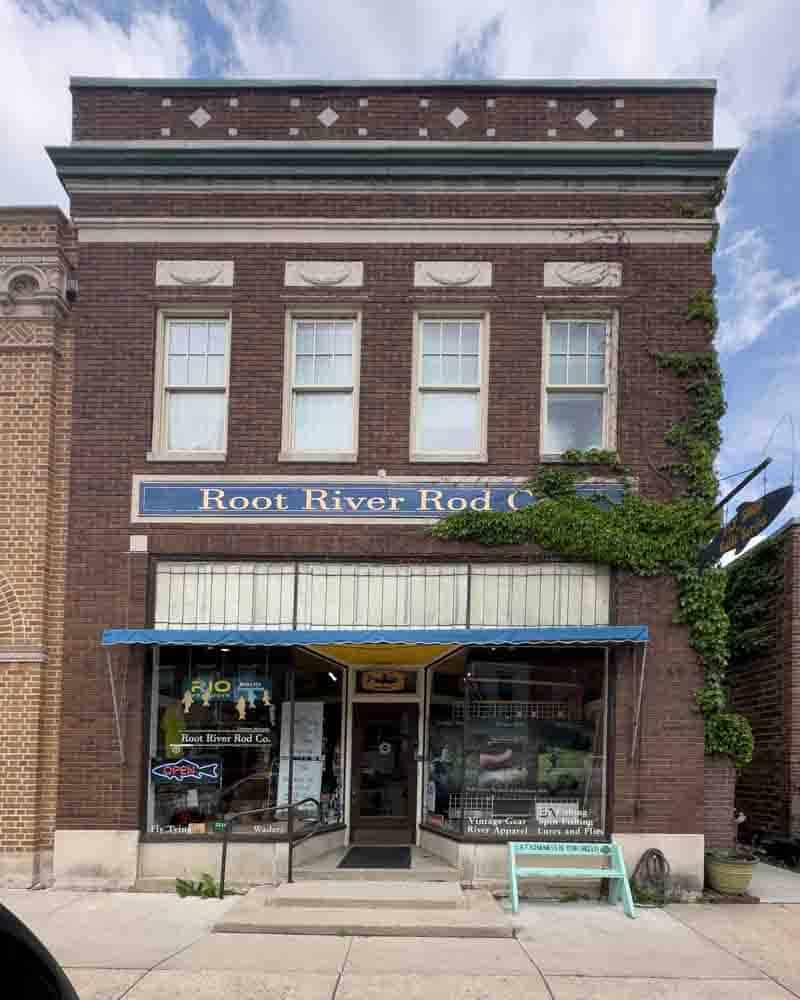 Vintage brick building with a storefront for Root River Rod Co. featuring an awning, signage, and ivy climbing the right wall, embodying classic small-town charm and fishing gear retail.