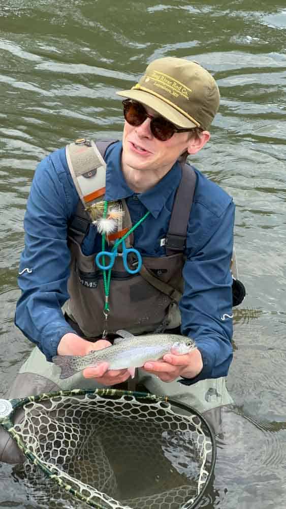 A woman fishing in a river holding a rainbow trout with a fishing net in the water, showcasing outdoor recreational fishing activities for seniors and nature lovers.