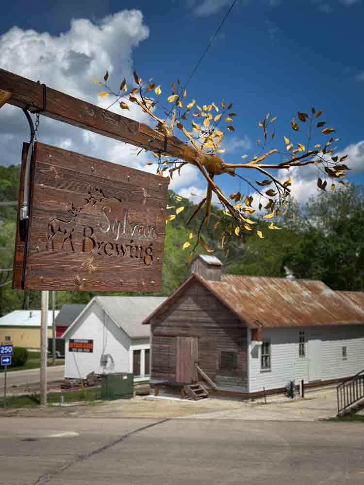Faded wooden sign for Sycamore Valley Tn Brewing hanging outside rustic brewery, vintage buildings, small town atmosphere, scenic rural backdrop, charming local craft beer spot, Old-fashioned outdoor storefront.