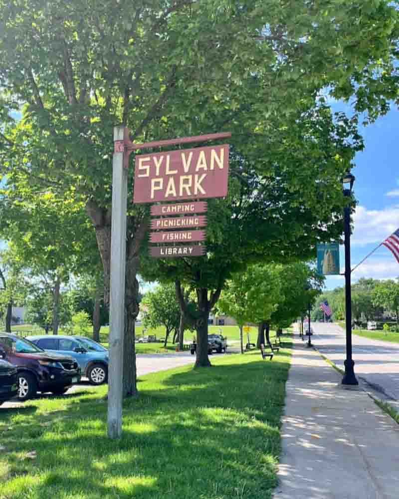 Sylvan Park sign with activities including camping, picnicking, fishing, and visiting the library, located in a lush green community park, perfect for outdoor family fun and relaxation.