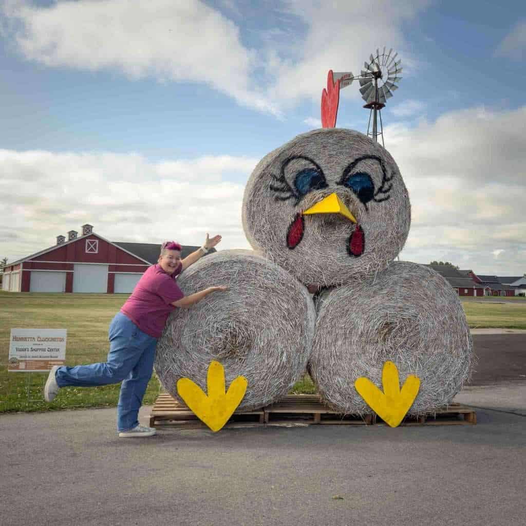 Colorful woman with pink hair at a farm fair posing with a giant hay bale chicken sculpture. The hay sculpture has a painted face, yellow beak, and large yellow feet, with a windmill on its head.