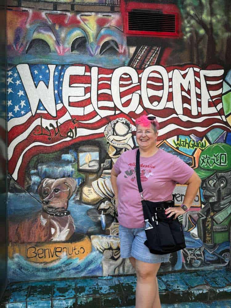 Vibrant graffiti mural welcoming visitors in a colorful urban setting with a cheerful grandma standing in front, highlighting community, travel, and adventurous spirit.