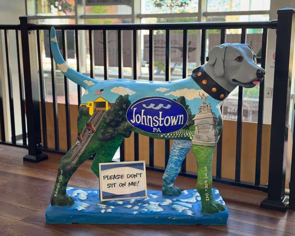 Colorful dog-shaped sculpture representing Johnstown PA with local landmarks and scenery, displayed indoors on wooden flooring; features humorous sign and vibrant artwork showcasing local pride.