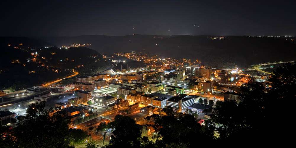 Brightly lit cityscape at night with illuminated buildings and streets, captured from an elevated viewpoint, showcasing urban life and nightlife in a scenic mountain setting.