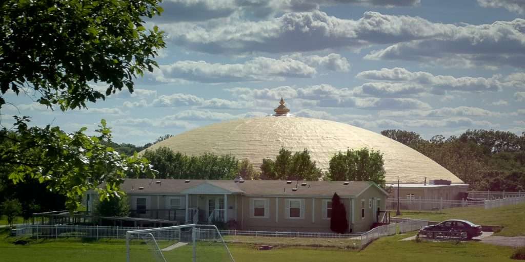 The Maharishi Golden Domes in Fairfield, Iowa rising behind trees and a row of nearby homes under a bright blue sky.